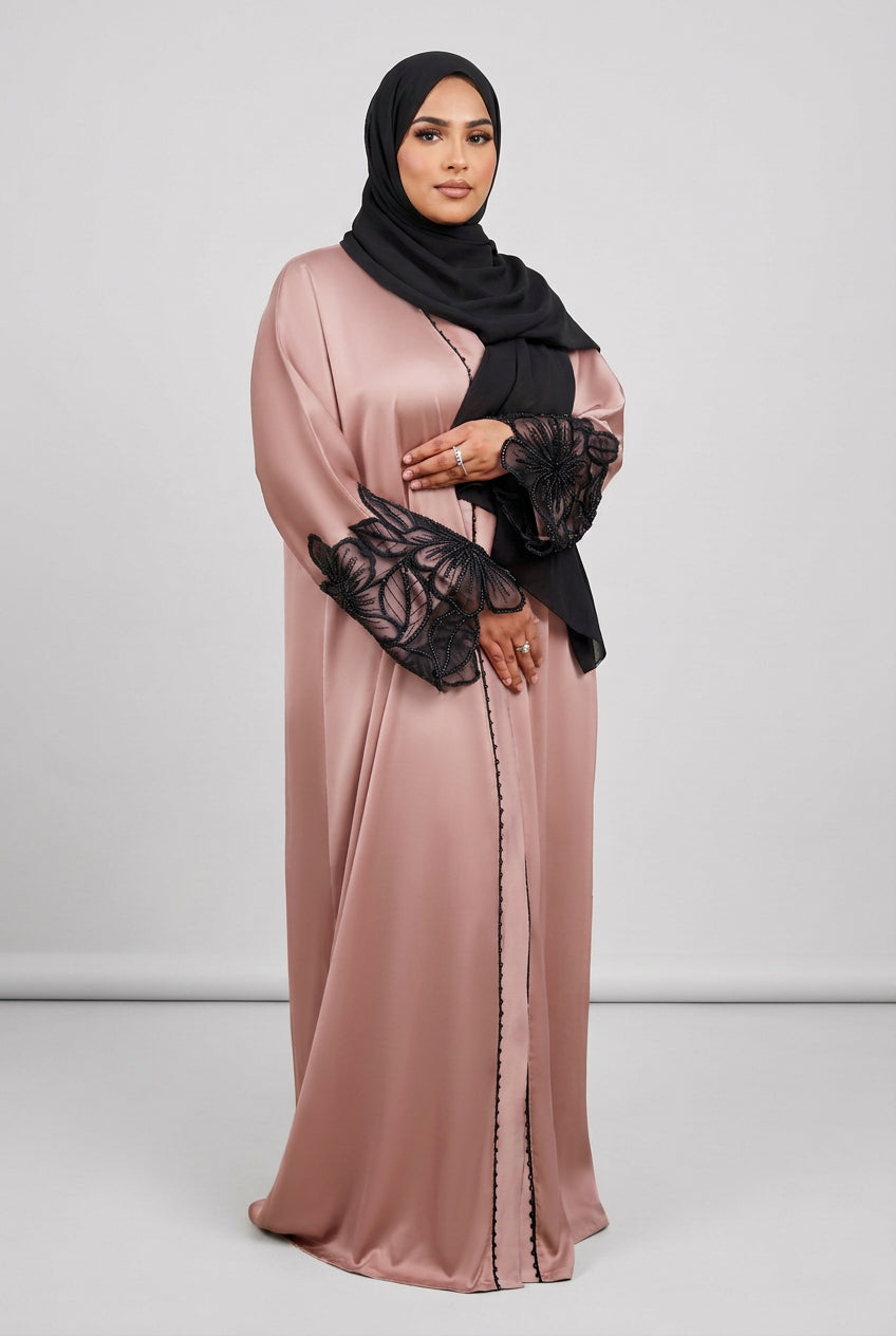 A woman wearing a long dusty blush pink abaya with black lace bell sleeves and a black hijab